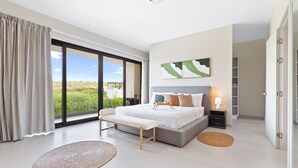 Ein modernes Schlafzimmer mit einem großen Bett, einer Bank, einem Nachttisch mit Lampe und Blick auf Grün durch das Fenster.