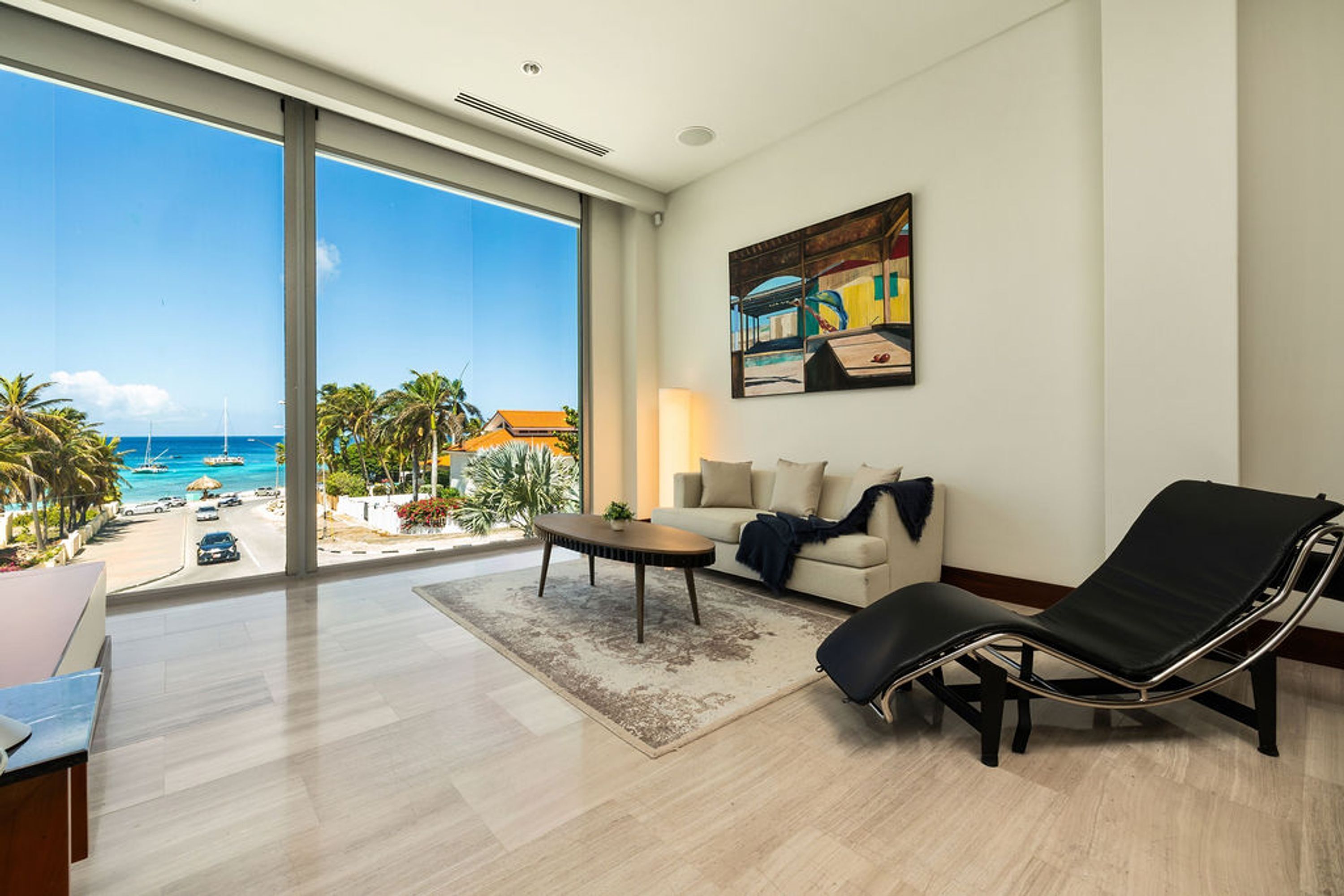 Un salón amplio con un gran ventanal con vistas a la playa, un sillón negro, un sofá color beige y una mesa de centro.
