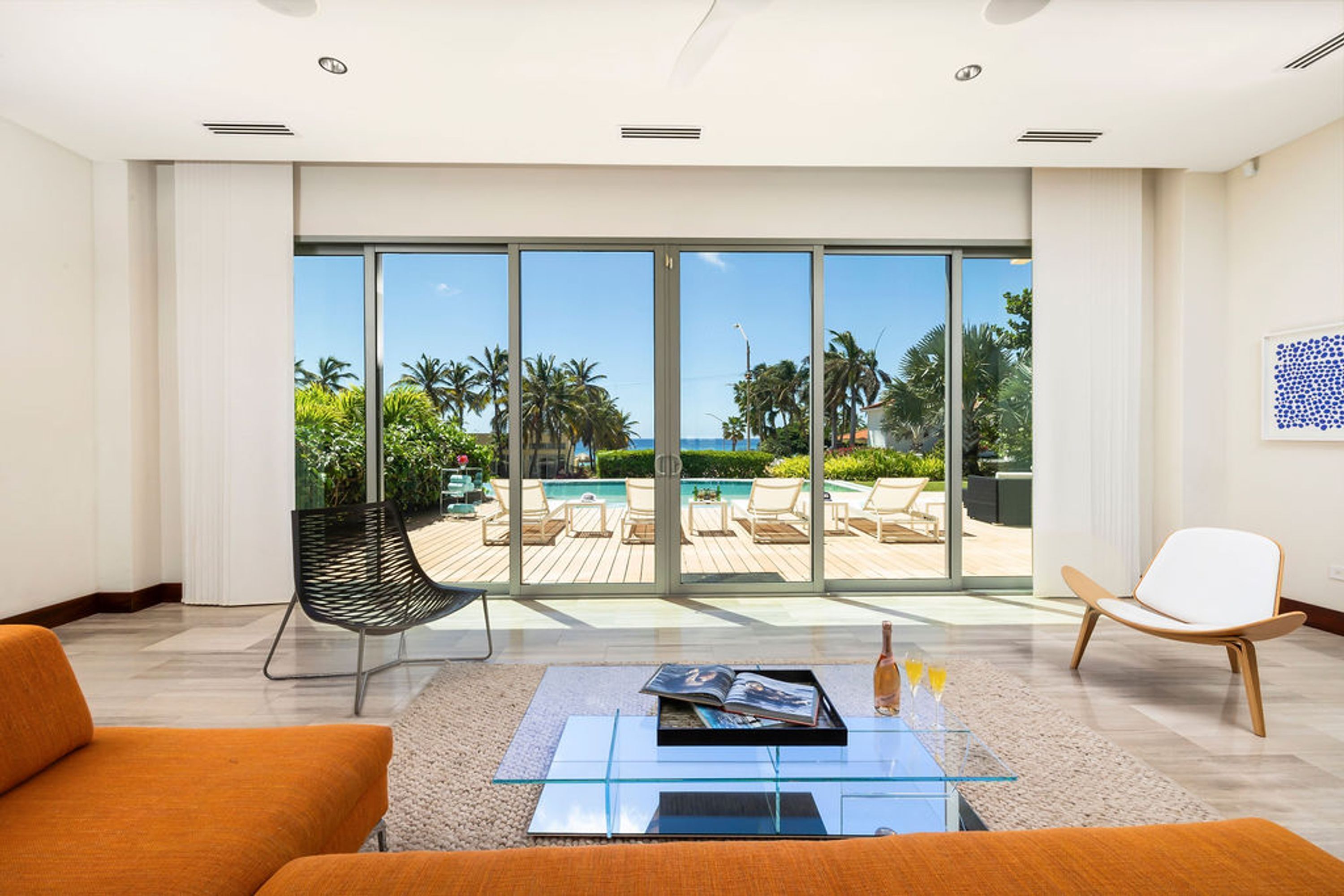 Una sala de estar moderna con una mesa de centro de vidrio, dos sofás naranja, una silla negra y una silla blanca. Grandes puertas corredizas de vidrio dan a un área de piscina exterior con sillones de descanso.