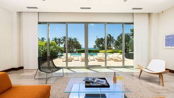 A modern living room with a glass coffee table, two orange sofas, a black chair, and a white chair. Large sliding glass doors lead to an outdoor pool area with lounge chairs.