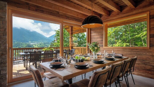 A wooden dining area with a table set for six, large windows, and a view of mountains. - Chalet Emeraude des Alpes - peaceful break in the mountains - OVO Network (Saint-Pierre-d'Entremont)