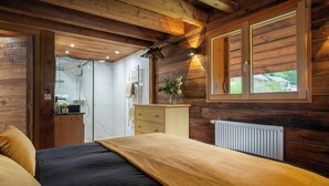 A bedroom with a wooden ceiling, a bed with a dark and light-colored blanket, a dresser, a radiator, and a window with a view of greenery outside. - Chalet Emeraude des Alpes - peaceful break in the mountains - OVO Network (Saint-Pierre-d'Entremont)
