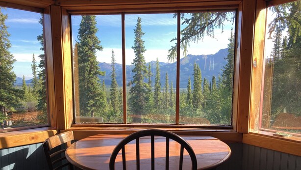 A corner room with a large window overlooking a forest and mountains. There is a wooden table with chairs and a small trash bin.