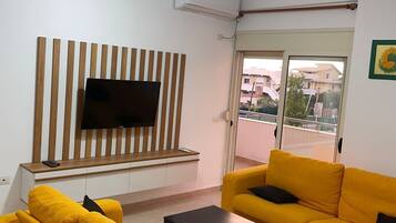 A living room with yellow sofas, a flat-screen TV on a wooden panel, a ceiling fan, and a view through a glass door.