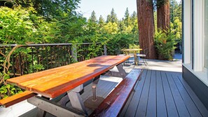 An outdoor wooden deck with picnic tables and benches, surrounded by lush greenery and tall trees.