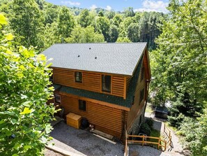 A two-story log cabin with a green roof, surrounded by dense forest. - Hot Tub, Fiber-optic WiFi, Pool Table, Playground, EV charger. Cozy Log Cabin. (Waynesville)