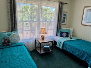 Ein Zimmer mit zwei Betten, einem kleinen Tisch mit Lampe, einem Teddybären und einem Fenster mit Vorhängen.