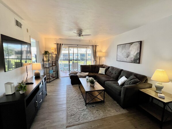 A living room with a sofa, coffee table, and a TV on a stand. There is a balcony with a view of the outside.