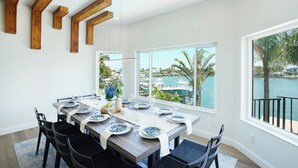 A dining area with a long table set for six, large windows with a view of the water, and wooden beams on the ceiling.
