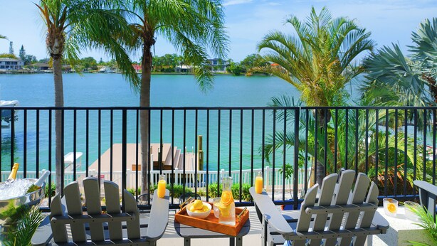 A balcony with two Adirondack chairs, a wooden tray with drinks, and a view of a body of water with palm trees.