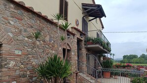 Ein Steinhaus mit einer Hausnummer, einer Treppe, die zu einem Balkon mit Topfpflanzen führt, und einem Eingang mit Blumen und einem Tor.