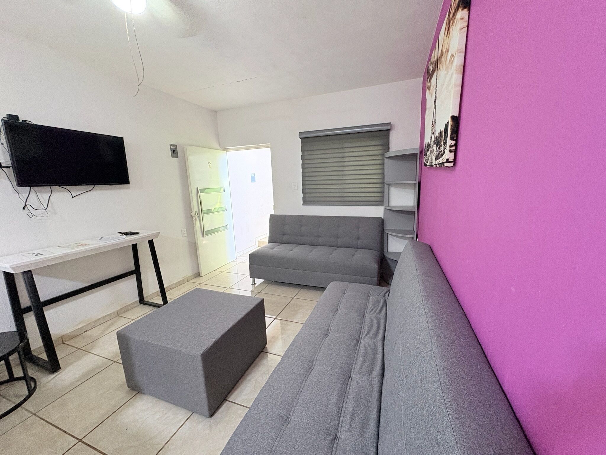 Une pièce avec un canapé gris, un petit pouf gris, un bureau blanc avec un téléviseur à écran plat et un mur rose orné d’un tableau encadré.