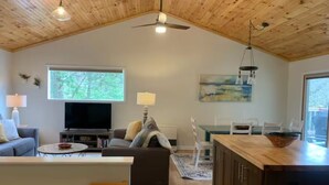 A spacious living area with a wooden ceiling, a large window, a television, a sofa, a dining table, and a kitchen area. - Howard's Creek Hideaway - Cozy mountain home close to downtown Boone & App State (Boone)