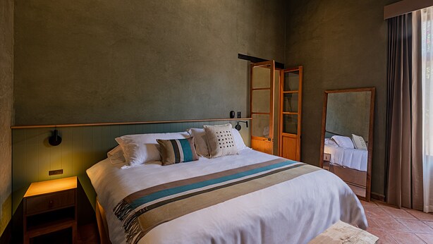 Un letto rifatto con cura, una coperta a righe, un comodino con una lampada e un grande specchio appeso al muro.