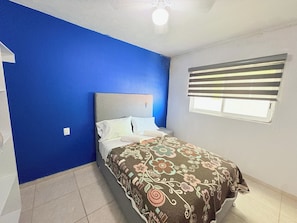 Un dormitorio con cama, paredes azules, ventana con persianas y ventilador de techo.