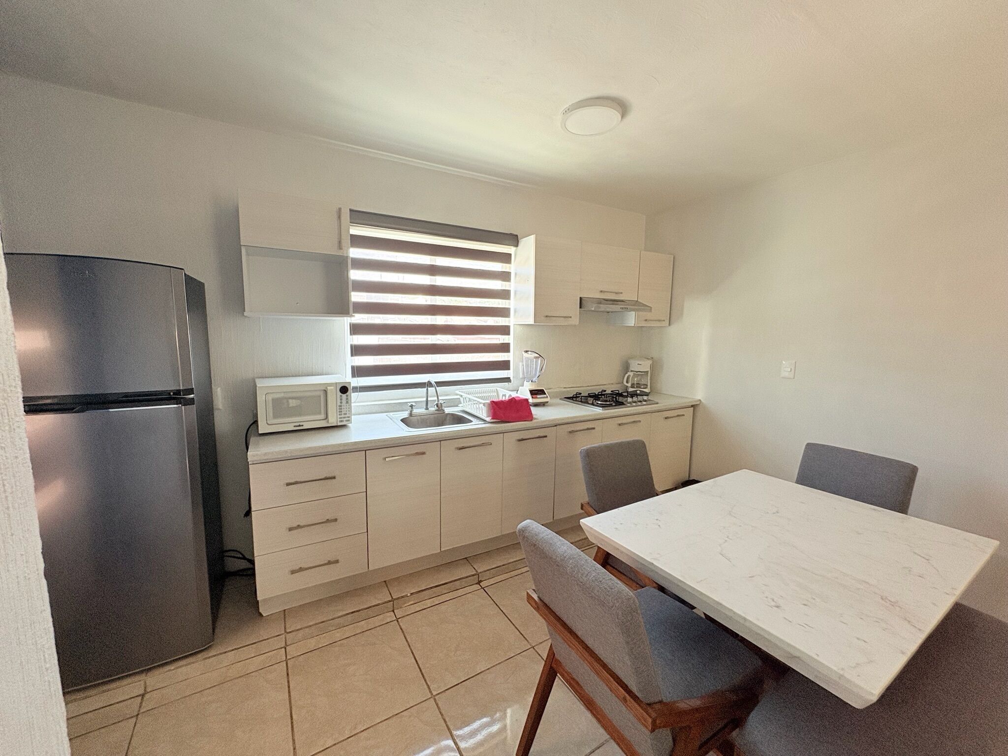 A compact kitchen with a refrigerator, microwave, sink, and stove. There is a dining table with chairs and a window with blinds.