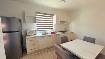 A compact kitchen with a refrigerator, microwave, sink, and stove. There is a dining table with chairs and a window with blinds.