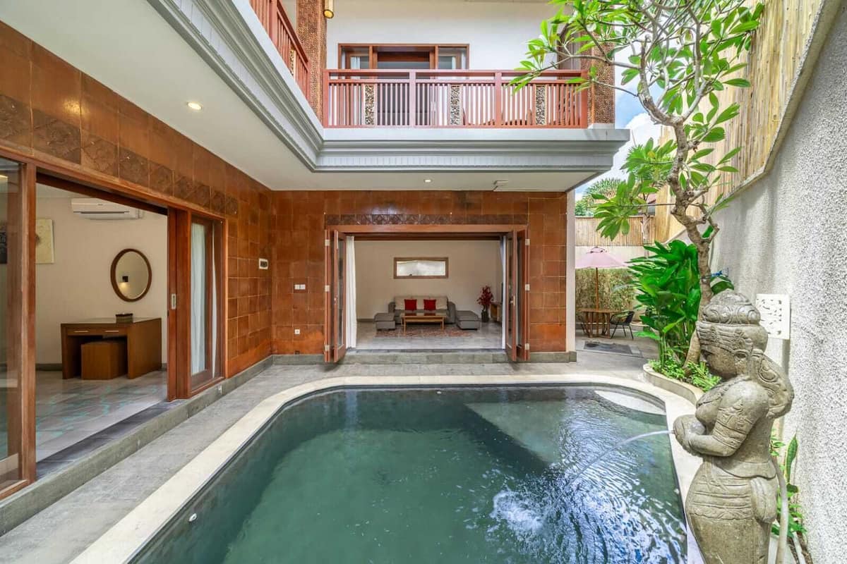 Une vaste piscine intérieure avec une zone de pont en bois, un coin salon comprenant un canapé et une table, et une statue située à droite.