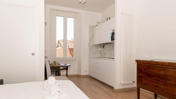 Un dormitorio con una cama, un aparador de madera, una cocina con armarios blancos y una ventana con vistas a edificios.