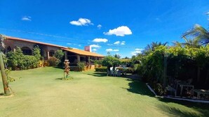 A courtyard with a building featuring a red-tiled roof and arched windows, surrounded by lush greenery and a clear blue sky. - Pousada Sol Nascente (Beberibe)