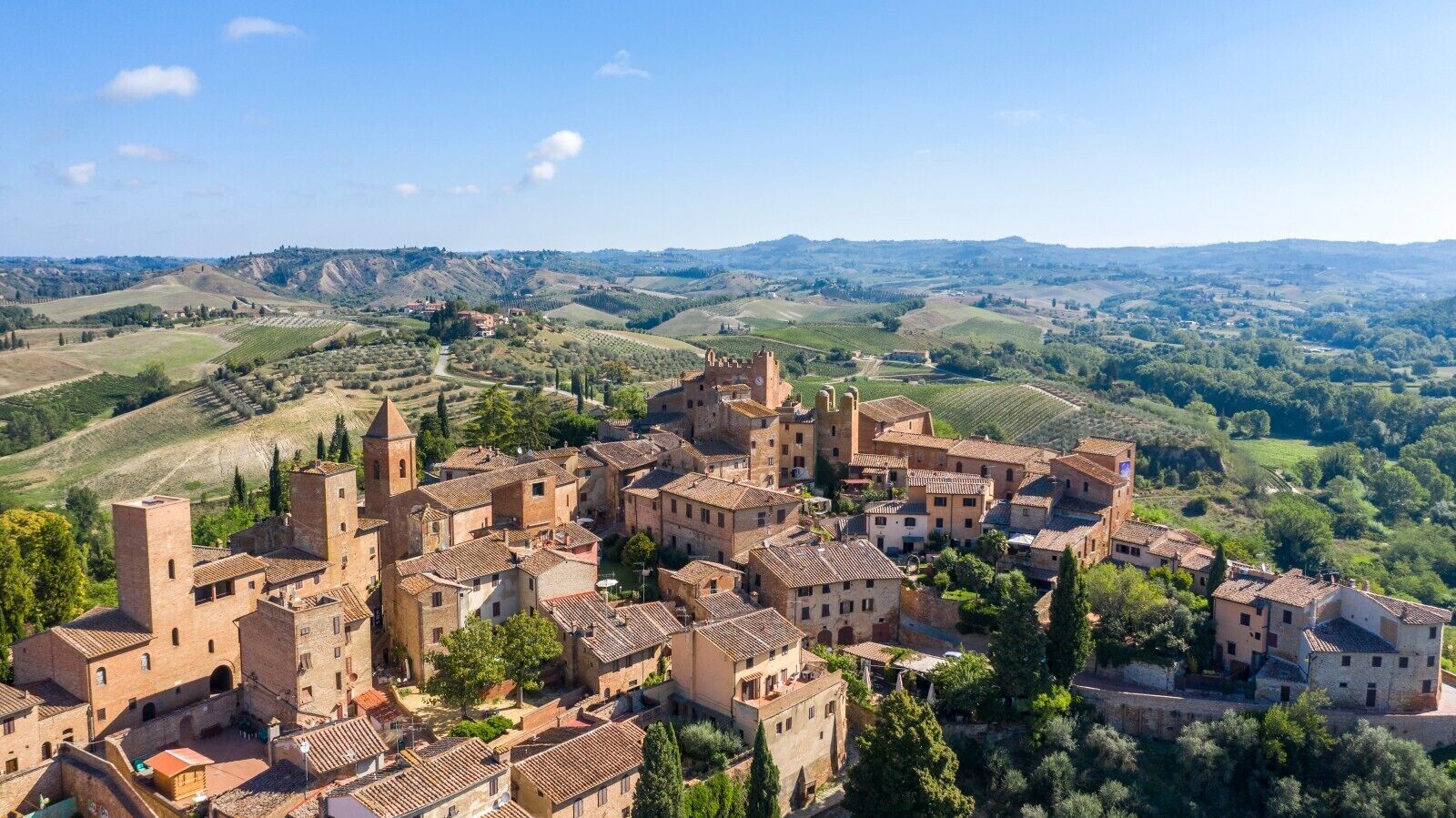 A historic town with medieval architecture, including towers and fortified walls, surrounded by rolling hills and vineyards.