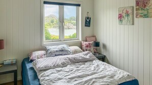 A bedroom with a bed, a nightstand, a lamp, a window with a view of greenery and a building, and wall decorations. - Cozy home in Hellvik with WiFi (Hellvik)