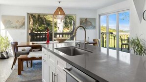 Une cuisine moderne dotée d’un grand îlot central, d’un coin repas avec une table et des chaises en bois, et offrant une vue sur une terrasse et un espace verdoyant grâce à de grandes fenêtres.