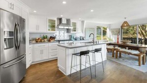 Une cuisine moderne avec des meubles blancs, un réfrigérateur en acier inoxydable, un îlot central avec des tabourets et une vue sur l’extérieur.