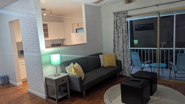 A living room with a sofa, a side table with a lamp, a small ottoman, and a balcony with chairs.