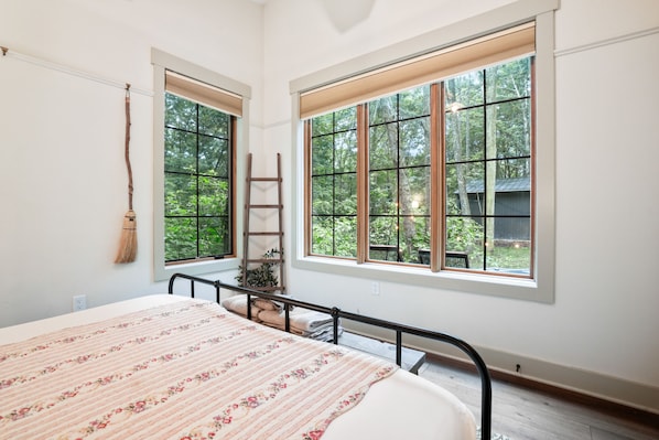 A bedroom with a large bed, two windows with wooden frames, a wooden ladder, and a broom hanging on the wall. - Evermore Cottage | Hocking Hills, Couples Retreat (Creola)