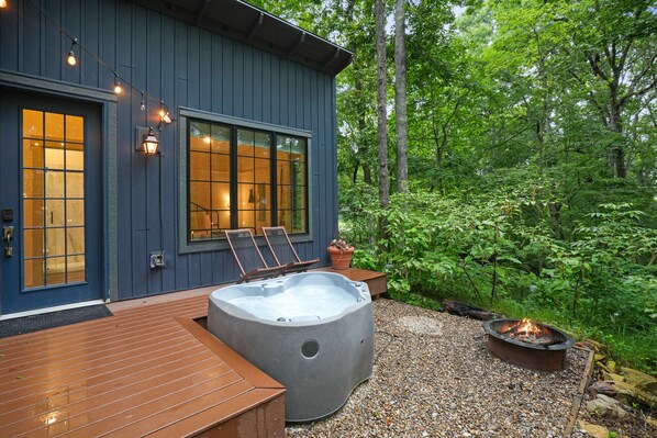 A wooden deck with a hot tub and a fire pit, surrounded by lush greenery.