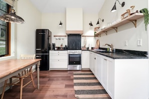 A modern kitchen with white cabinets, a black refrigerator, a stainless steel oven, and a wooden dining table. - Evermore Cottage | Hocking Hills, Couples Retreat (Creola)