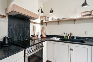 Eine moderne Küche mit weißen Schränken, einer schwarzen Arbeitsplatte, einem Herd und einem Spülbecken. Zwei Wandleuchten, ein Holzregal mit Geschirr und eine hängende Pflanze sind ebenfalls vorhanden.