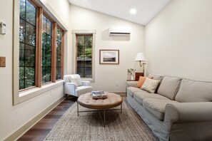 A spacious living room with large windows, a beige sofa, a round coffee table, and a striped armchair.