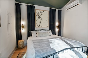 Un dormitorio moderno con cama, cortinas oscuras, aire acondicionado de pared y un taburete de madera.