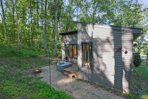 Una cabaña moderna con terraza, jacuzzi y una fogata en un área boscosa.