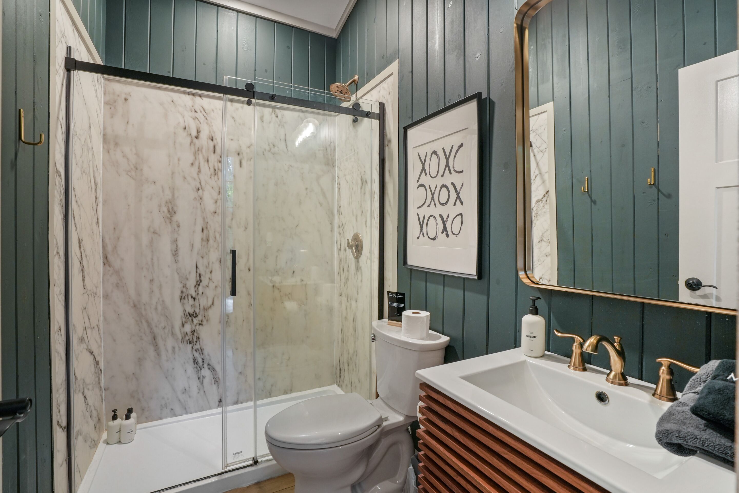 A bathroom with a glass-enclosed shower, a toilet, a sink with gold faucets, and a framed sign on the wall.
