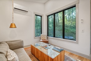 Un salon moderne avec un canapé, une table basse sur laquelle sont posés des livres, une lampe suspendue et de grandes fenêtres donnant sur une forêt.