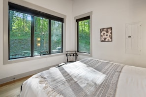 Un dormitorio con ventanas amplias, una cama con manta a rayas, una mesita pequeña y un cuadro enmarcado en la pared.