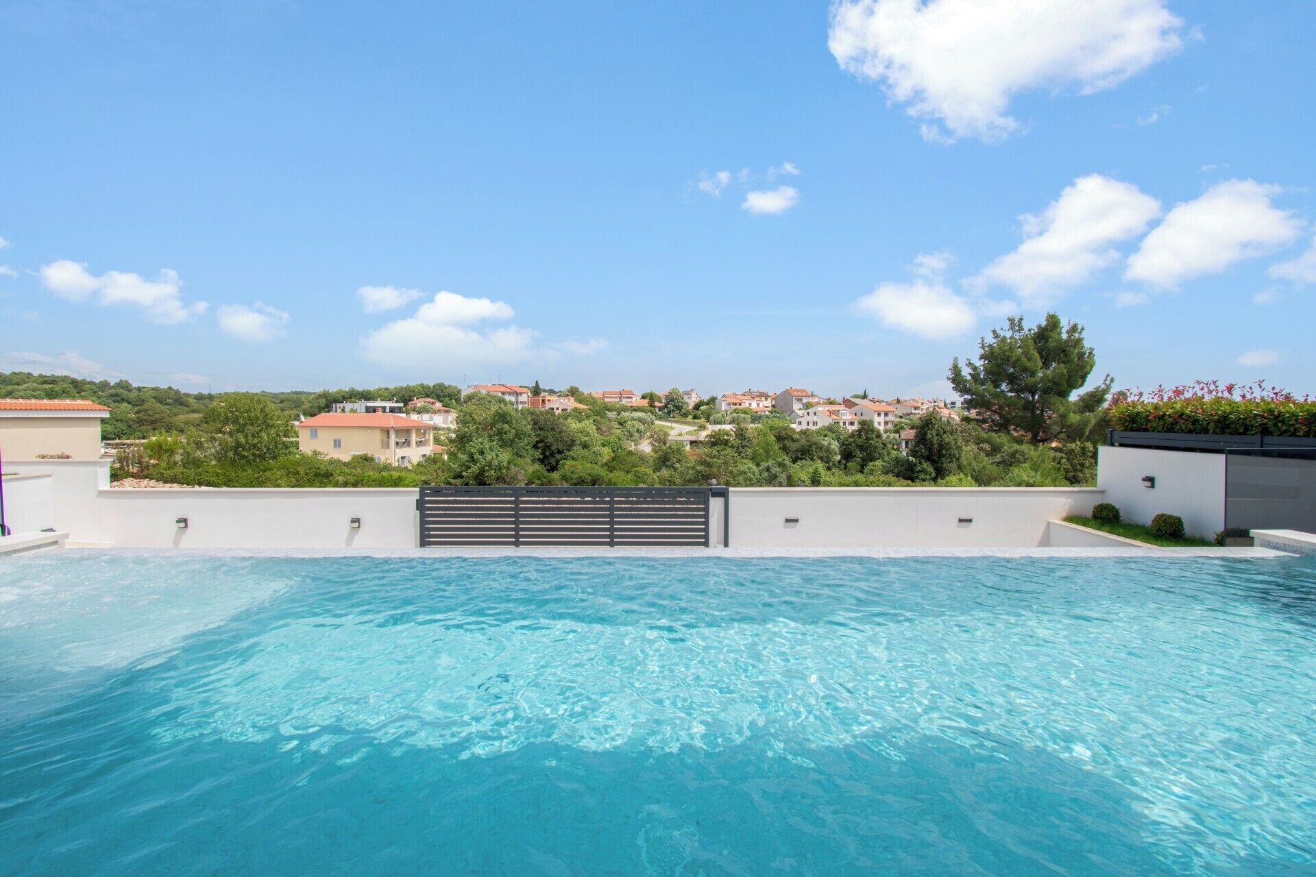 Una piscina con acqua cristallina e blu, circondata da un muro bianco e una recinzione nera. Sullo sfondo, si vedono case e alberi sotto un cielo sereno.
