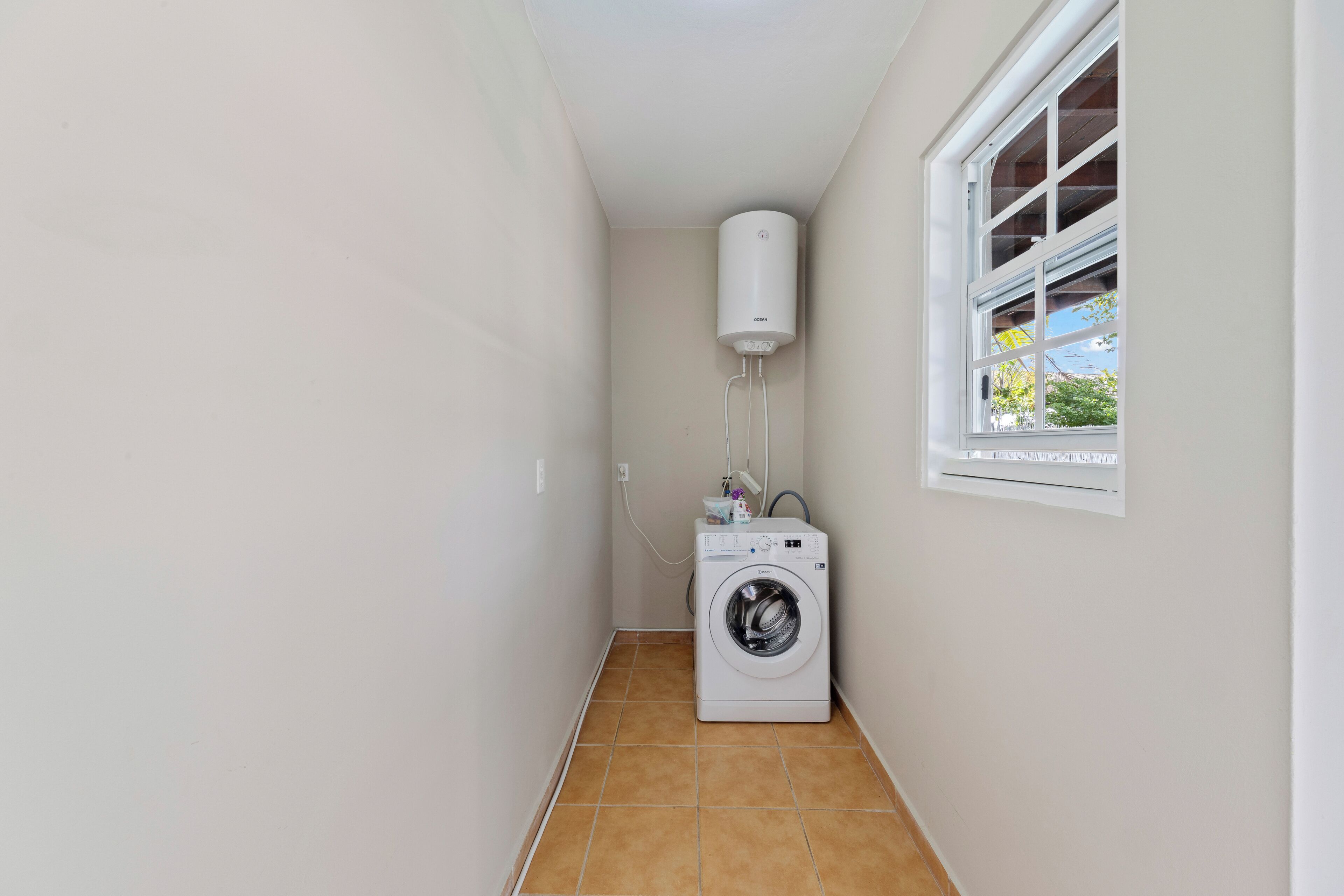 A laundry room with a washing machine, a water heater, and a window with a view of greenery outside.