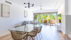 A modern living room with a glass dining table, beige chairs, a sofa, and a large window offering a view of greenery. - Ref. 421409 (Estepona)
