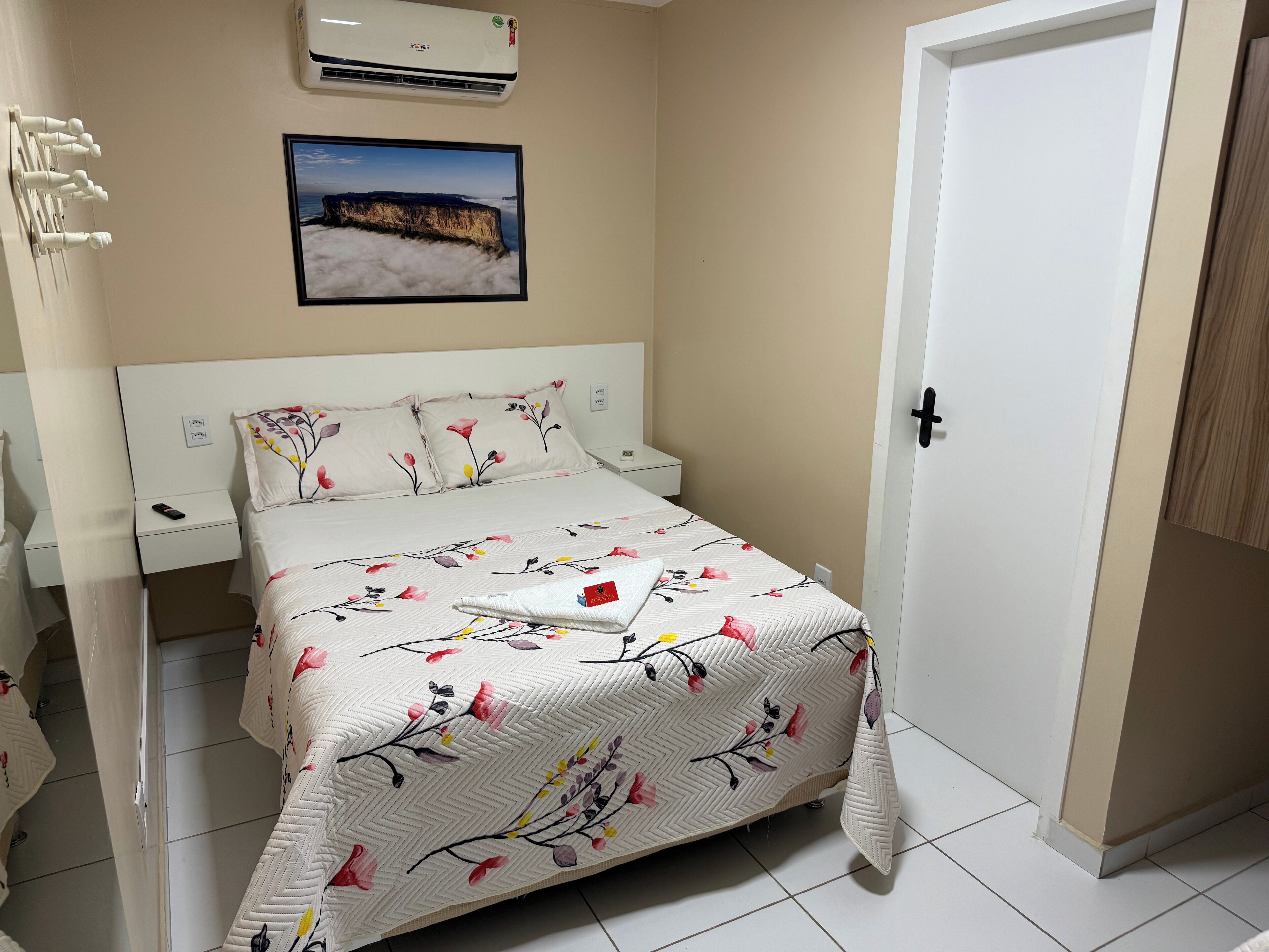 Une chambre à coucher comprenant un lit, une table de chevet, un climatiseur fixé au mur, un tableau encadré et une porte.