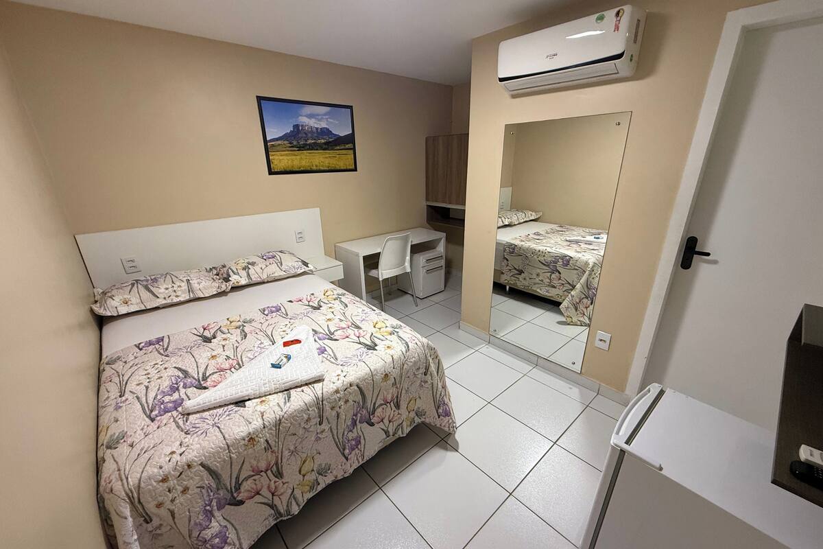 A bedroom with a bed, a desk, a chair, an air conditioner, and a mirror.