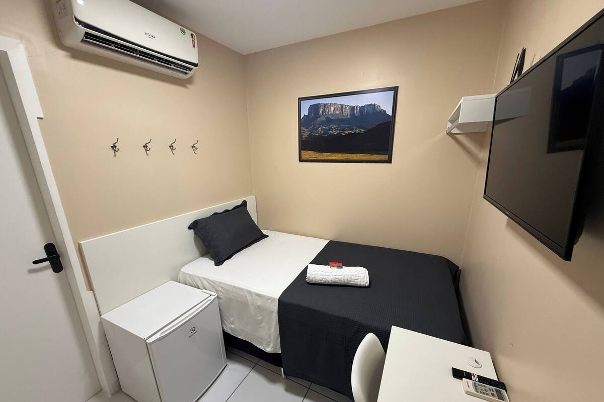 A compact hotel room with a bed, a small desk, a chair, a TV, an air conditioner, and a picture on the wall.