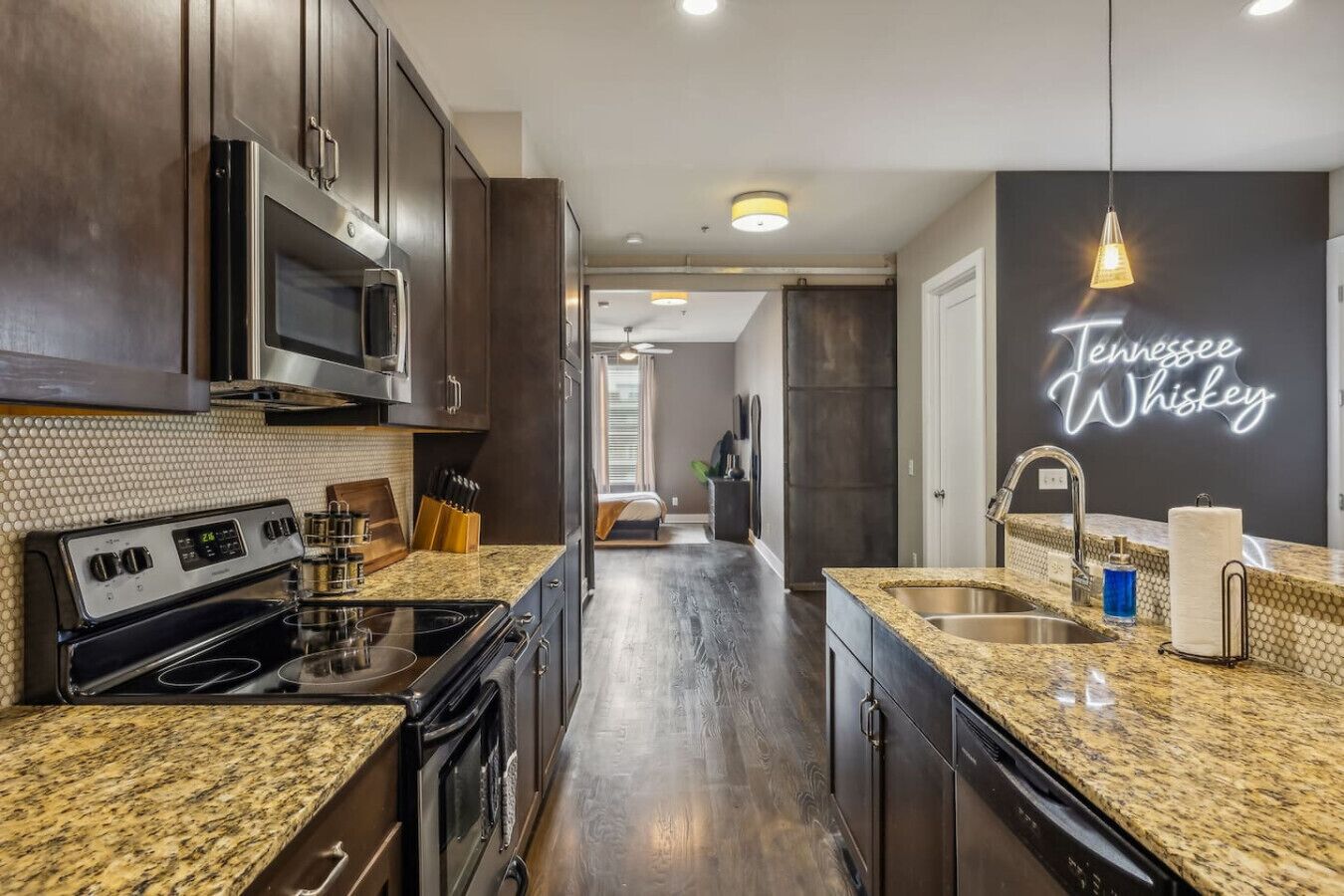 A modern kitchen with dark wood cabinets, granite countertops, stainless steel appliances, and a neon sign reading "Tennessee Whiskey".