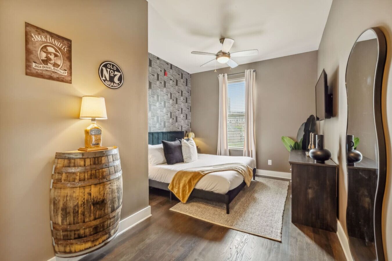 A bedroom with a bed, a ceiling fan, a wooden barrel, a lamp, a mirror, and a wall with a logo.
