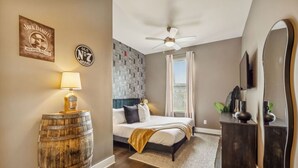 A bedroom with a bed, a ceiling fan, a wooden barrel, a lamp, a mirror, and a wall with a logo.