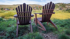 Two Adirondack chairs in a grassy field with a view of houses on a hill in the background. - Cozy Granby Escape: Ski, Hike & Relax (Granby)