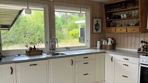 A kitchen with white cabinets, a wooden ceiling, a large window, and a built-in shelf with dishes. - Holiday home close to the beach in Nødebohuse (Hundested)